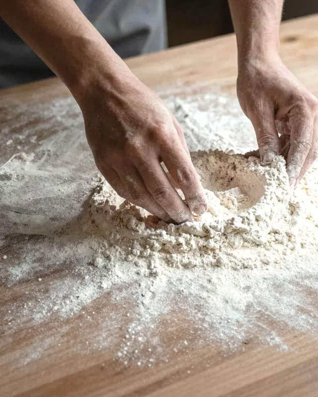 A baker's hands working flour across a wooden bench in soft window light.